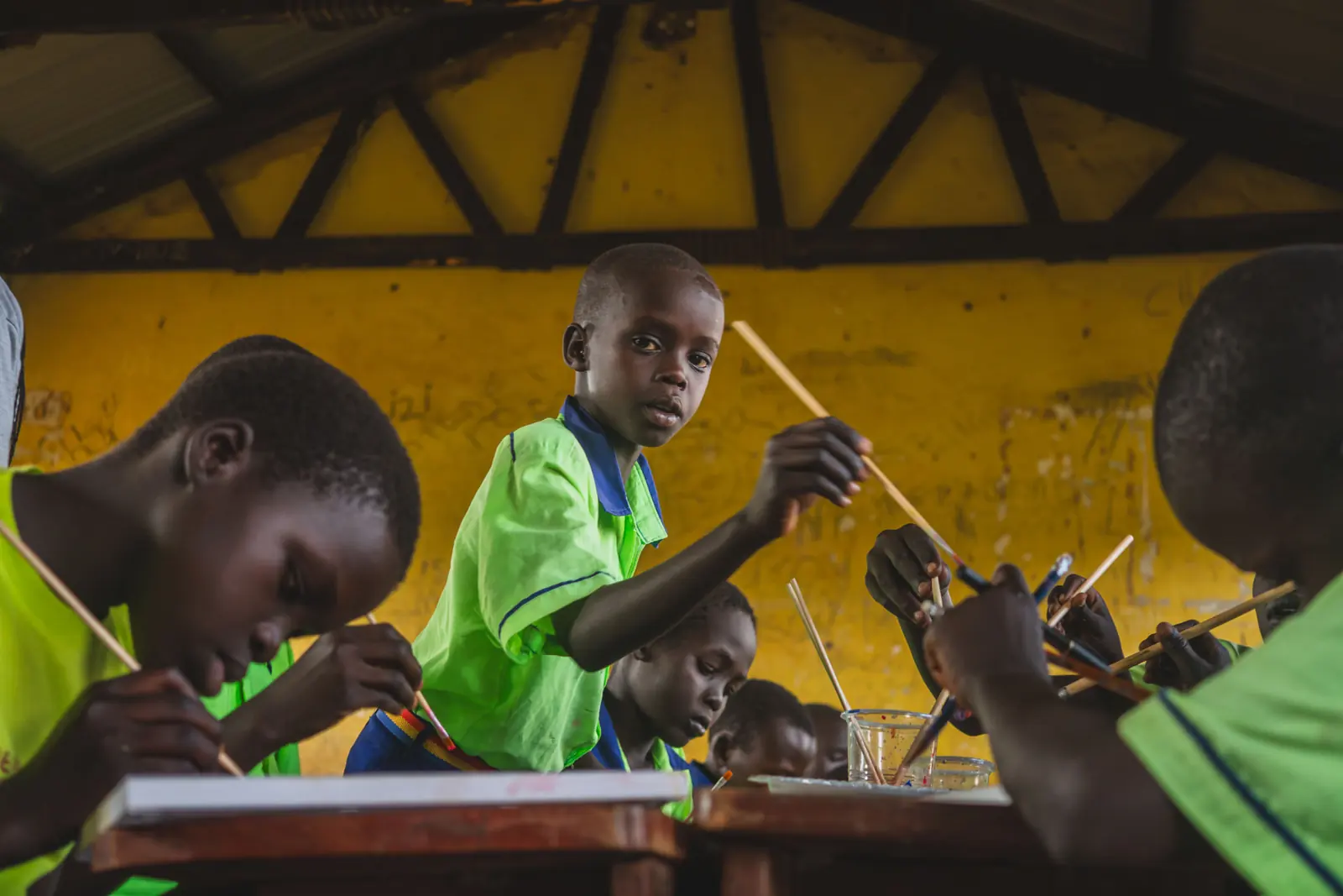 Children painting side by side at wooden desks in the Kampala art class